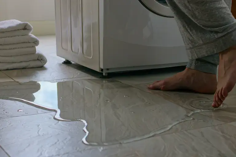 Water spreading across a laundry room floor from behind a washing machine